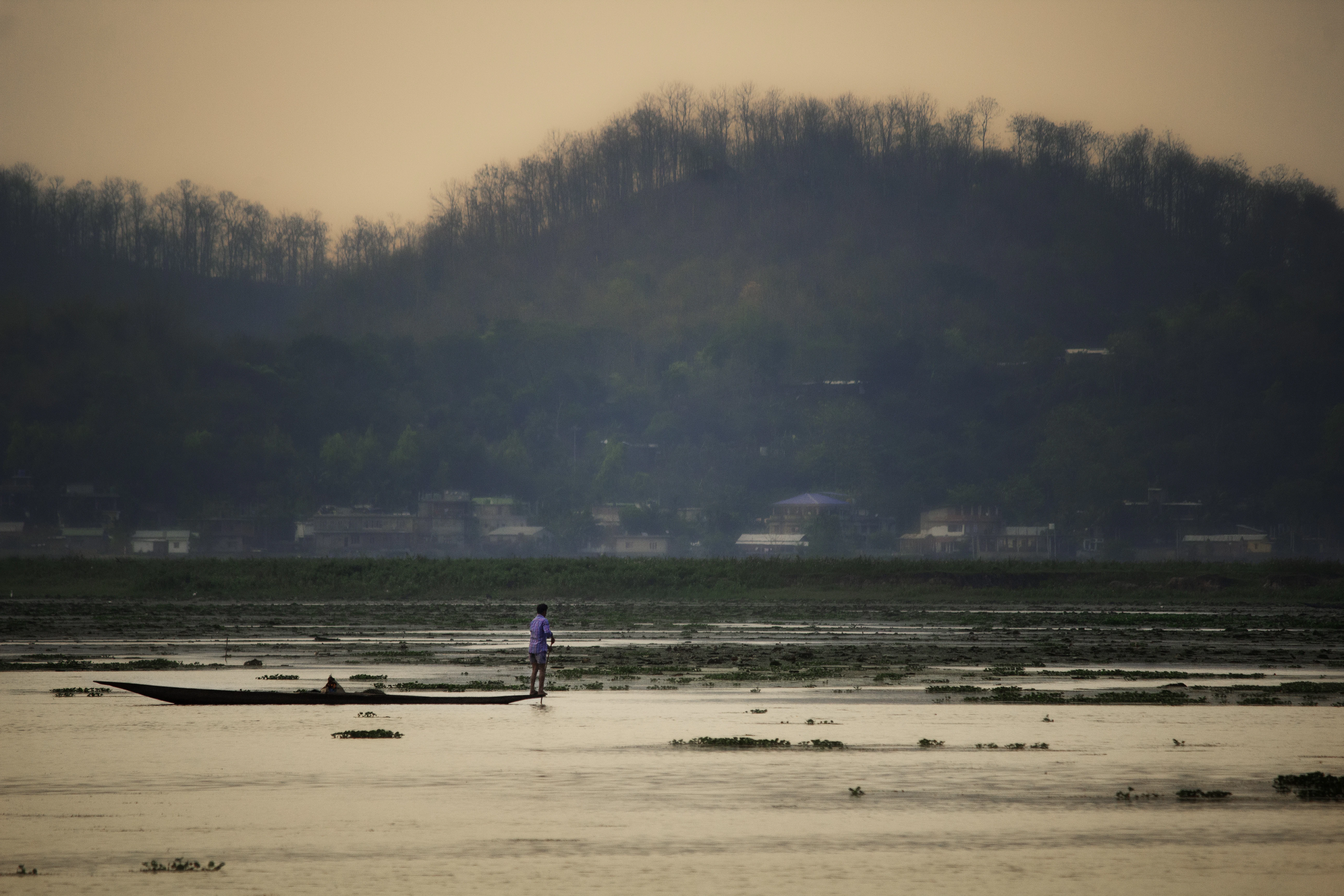 A lone figure poles a boat across a calm lake.