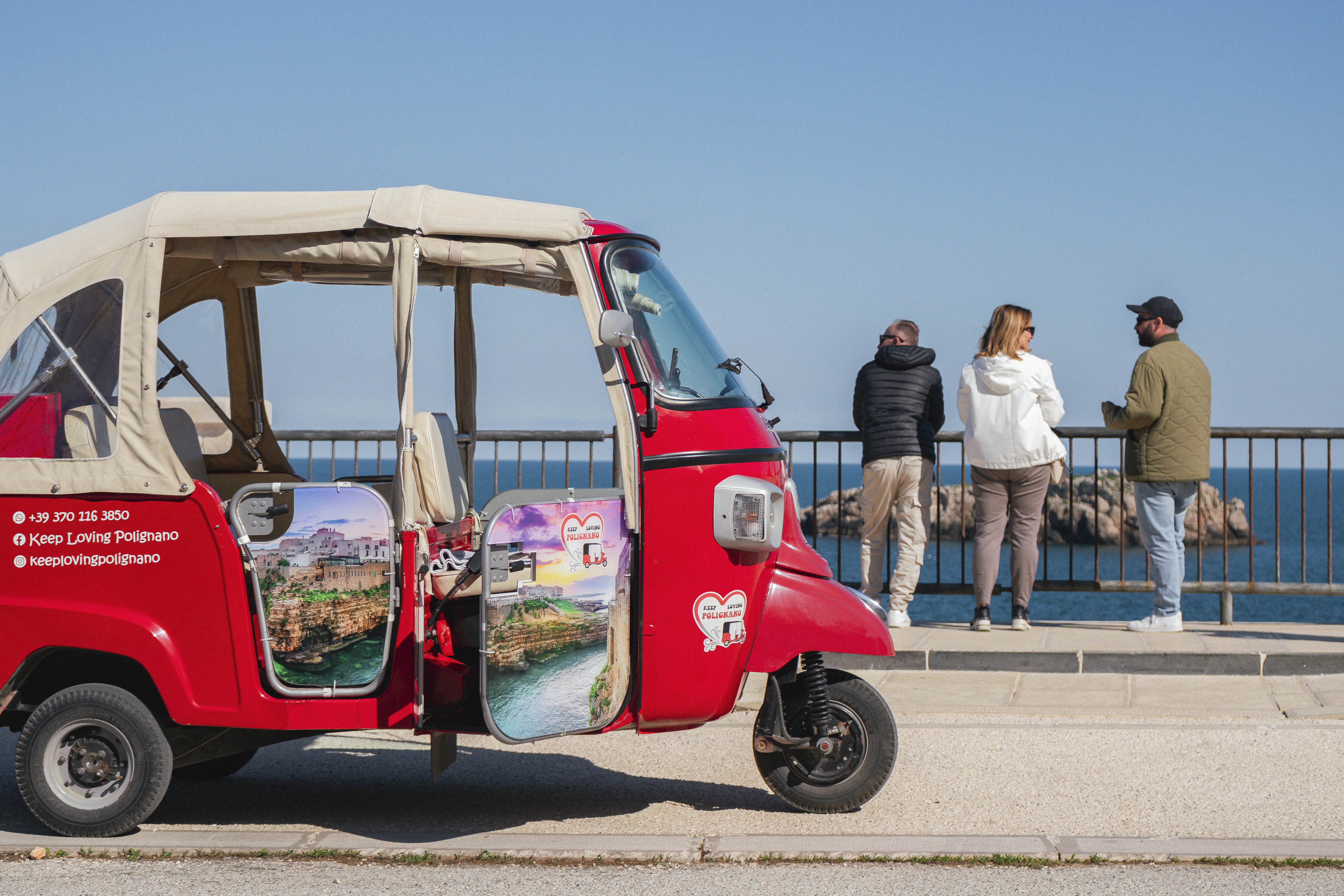 A red tuk-tuk awaits near tourists by the sea.